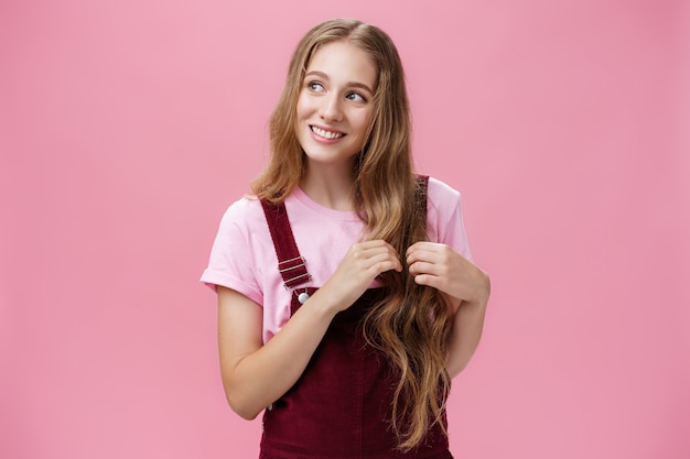 Tonto, amable y simpática chica encantadora que sueña despierta recogiendo mechones de cabello y mirando a la izquierda con una bonita sonrisa linda, imaginando una hermosa escena posando coqueta y feliz sobre un fondo rosa.