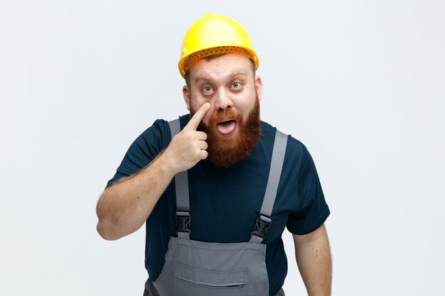 Tonto joven trabajador de la construcción con casco de seguridad y uniforme mirando a la cámara mostrando la lengua tirando de su párpado inferior hacia abajo aislado sobre fondo blanco.