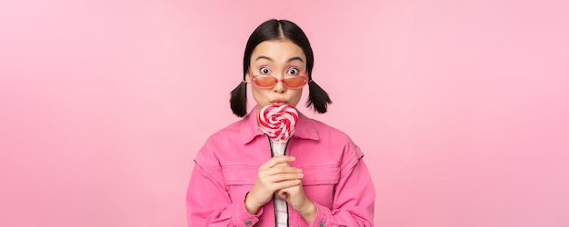 Tonto y lindo modelo femenino asiático lamiendo piruleta comiendo caramelos dulces y sonriente mirando emocionado de pie sobre fondo de color rosa