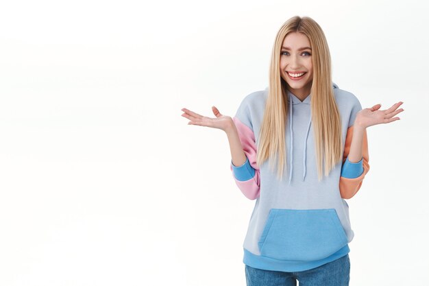 Tonto riendo linda chica rubia en sudadera con capucha, encogiéndose de hombros y extendiendo las manos hacia los lados, sonriendo como diciendo lo siento