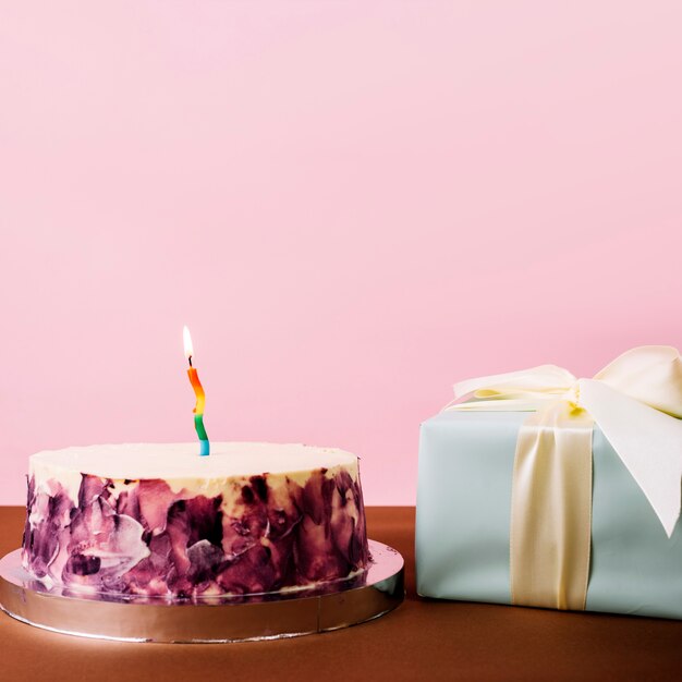 Torta deliciosa con la vela iluminada y la caja de regalo envuelta contra fondo rosado