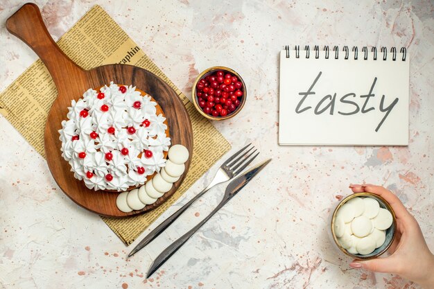 Torta de vista superior con crema pastelera blanca sobre tablero de madera en periódico. chocolate blanco en un tazón en la mano femenina. sabroso escrito en el cuaderno