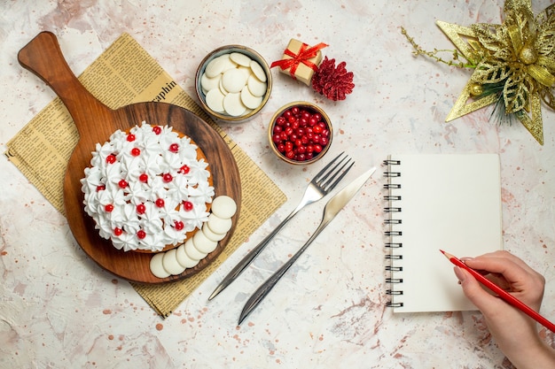 Torta de vista superior con crema pastelera en tablero de madera en periódico. ornamento de navidad, cuaderno y lápiz rojo en mano de mujer