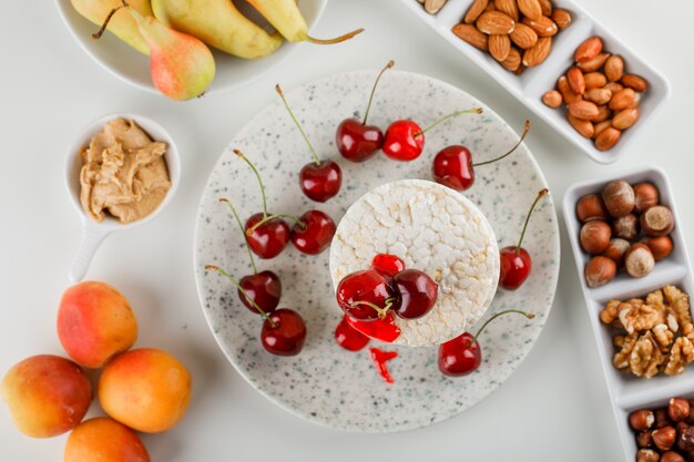 Tortas de arroz con cereza, nueces, pera, albaricoque, mantequilla de maní en una plataforma sobre fondo blanco, vista superior.