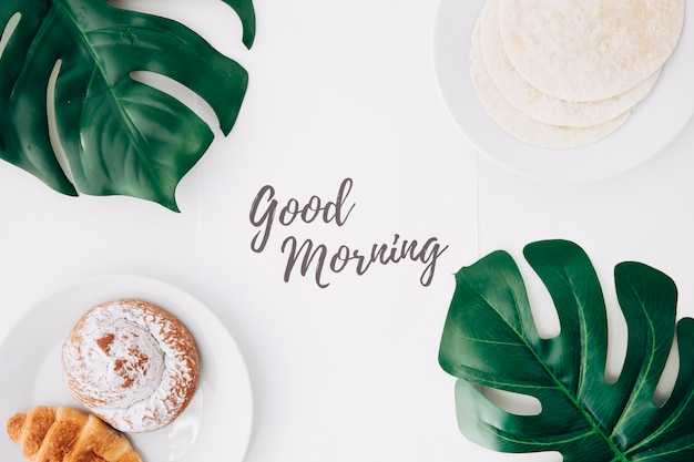 Tortillas de harina fresca; bollo al horno Desayuno de croissant con texto de buena mañana en papel y hojas de monstruo verde sobre fondo blanco