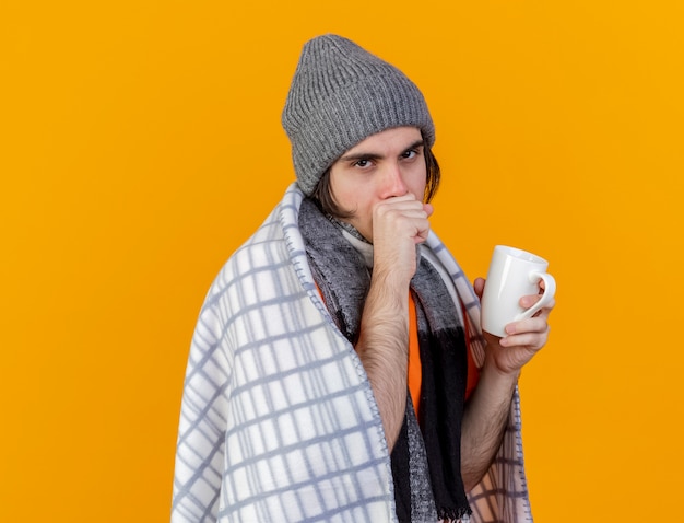 Tos joven enfermo con sombrero de invierno con bufanda envuelta en cuadros sosteniendo una taza de té aislado en naranja