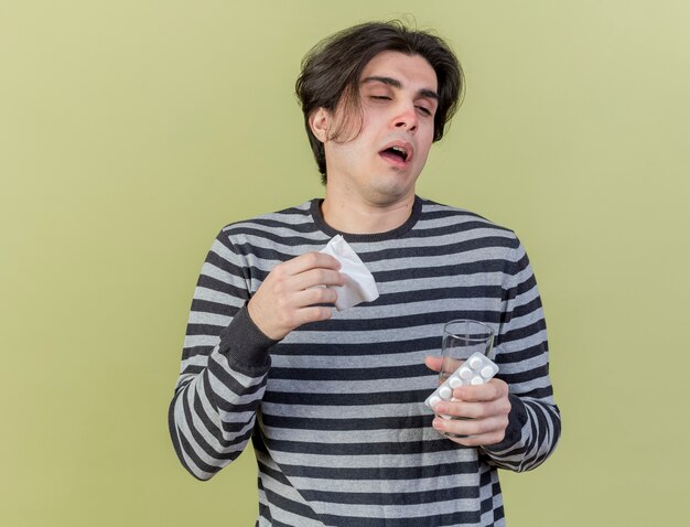 Tos joven enfermo sosteniendo una servilleta y un vaso de agua con pastillas aislado en verde oliva