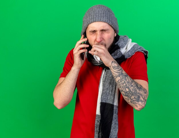 Toser joven enfermo caucásico vistiendo gorro y bufanda de invierno sostiene tejido hablando por teléfono