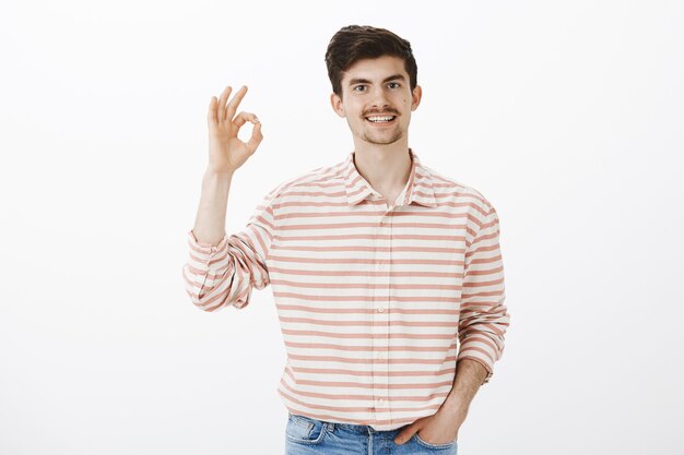 Totalmente de acuerdo contigo. Toma interior de un chico caucásico feliz tranquilo con barba y bigote, levantando las manos con un gesto aceptable, aprobando el concepto, pensando que la idea es excelente, de pie sobre una pared gris