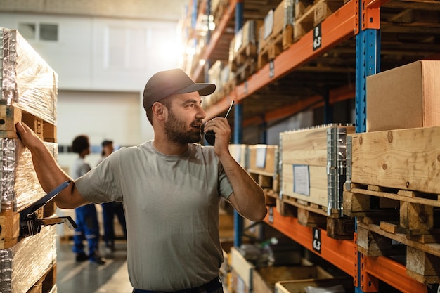 Foto gratuita trabajador de almacén masculino comunicándose con alguien a través de un walkietalkie mientras está de pie entre estantes en el almacén de distribución