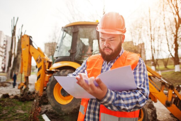 Trabajador de barba brutal traje de hombre trabajador de la construcción en casco naranja de seguridad contra traktor con papel de plan en las manos