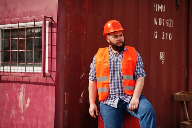 Trabajador de barba brutal traje de hombre trabajador de la construcción en casco naranja de seguridad sentado en barril rojo