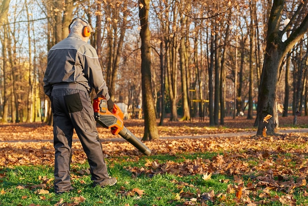 Foto gratuita trabajador de cabello gris usando soplador de hojas en el parque de la ciudad en octubre vista trasera de un hombre mayor enfocado