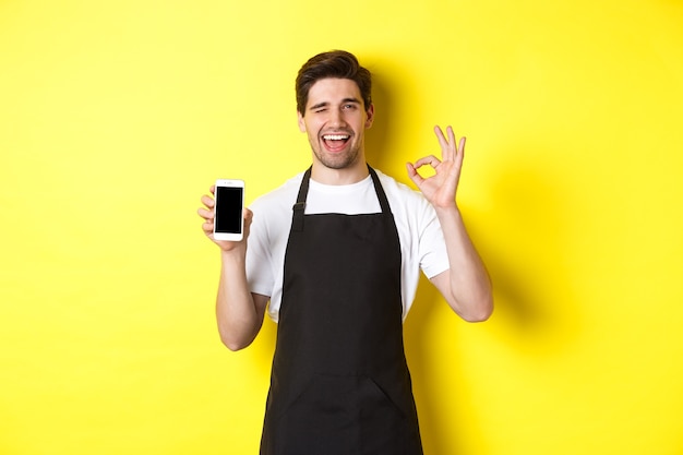 Trabajador de cafetería guapo mostrando signo ok y pantalla de teléfono inteligente, recomendando la aplicación, de pie sobre fondo amarillo.