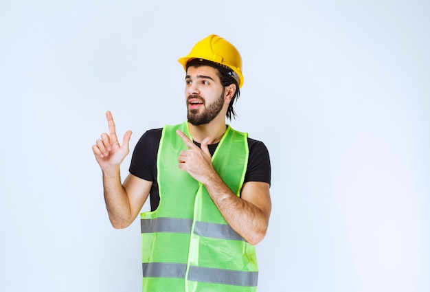 Trabajador en casco amarillo y chaqueta verde apuntando hacia arriba.