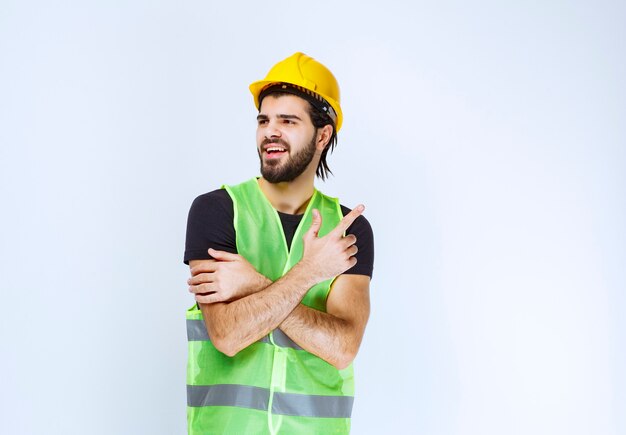 Trabajador en casco amarillo y chaqueta verde apuntando hacia arriba.