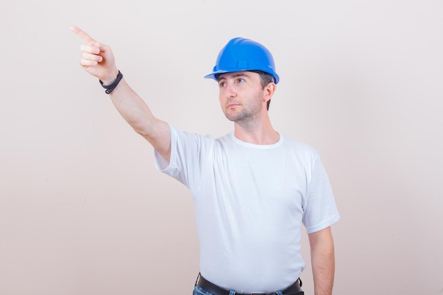 Trabajador de la construcción apuntando hacia afuera en camiseta, jeans, casco y mirando enfocado