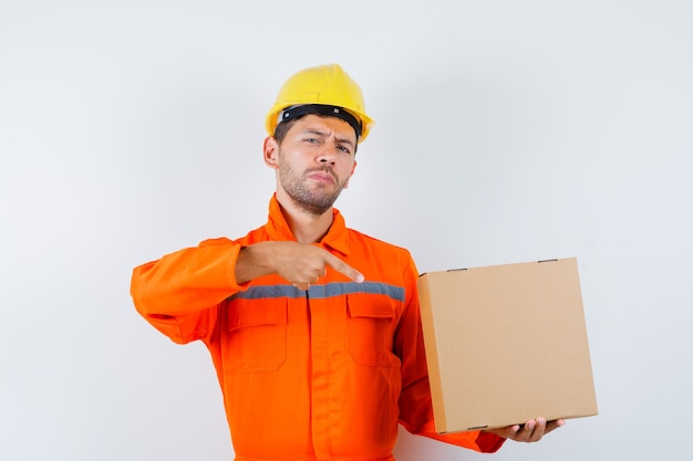 Trabajador de la construcción apuntando a la caja de cartón en uniforme, vista frontal del casco.