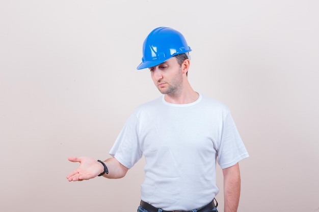 Trabajador de la construcción en camiseta, jeans, casco manteniendo la palma de la mano en forma interrogativa y con aspecto serio