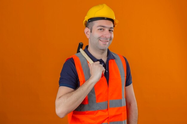 Trabajador de la construcción con chaleco y casco de seguridad amarillo con martillo con una sonrisa en la cara aislada en naranja