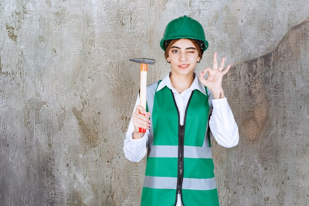 Trabajador de la construcción femenina en casco verde sosteniendo un martillo y dando el signo de ok