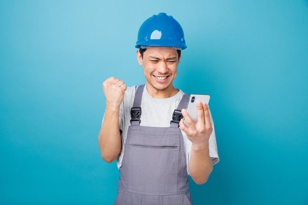 Trabajador de la construcción joven agresivo con casco de seguridad y uniforme sosteniendo y mirando el teléfono móvil haciendo gesto de sí