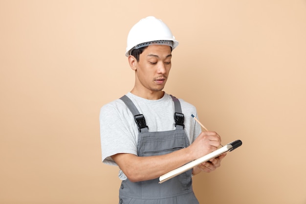 Trabajador de la construcción joven concentrado con casco de seguridad y uniforme escribiendo con lápiz en el bloc de notas