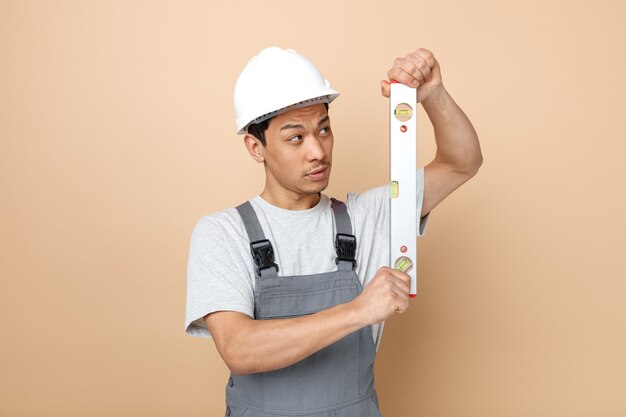 Trabajador de la construcción joven confundido con casco de seguridad y uniforme sosteniendo y mirando la regla de nivel