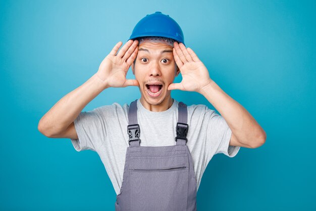 Trabajador de la construcción joven emocionado con casco de seguridad y uniforme manteniendo las manos en la cabeza