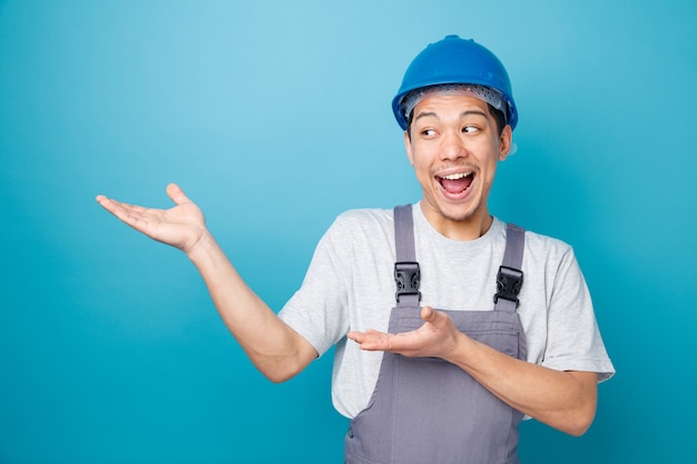 Trabajador de la construcción joven emocionado con casco de seguridad y uniforme mirando y apuntando al lado con las manos