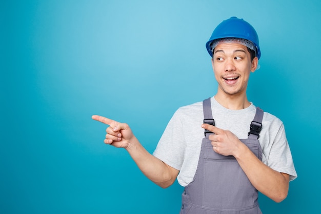 Trabajador de la construcción joven emocionado con casco de seguridad y uniforme mirando y apuntando a un lado