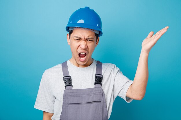 Trabajador de la construcción joven enojado con casco de seguridad y uniforme mostrando gritos de mano vacía