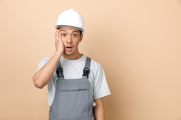 Trabajador de la construcción joven preocupado con casco de seguridad y uniforme manteniendo la mano en la cara