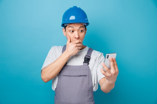 Trabajador de la construcción joven preocupado con casco de seguridad y uniforme sosteniendo y mirando el teléfono móvil manteniendo la mano en la boca