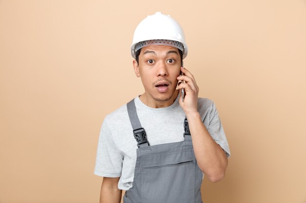 Trabajador de la construcción joven sorprendido con casco de seguridad y uniforme hablando por teléfono