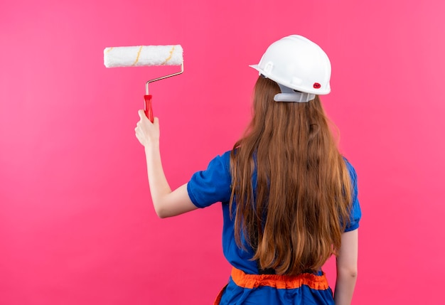 Trabajador de construcción joven en uniforme de construcción y casco de seguridad de pie con la espalda yendo a pintar con rodillo de pintura sobre pared rosa