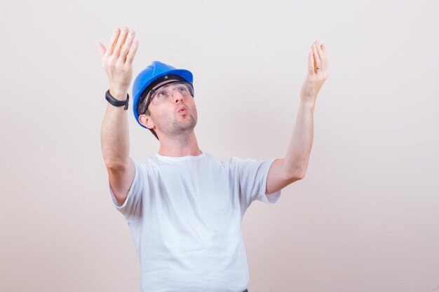 Trabajador de la construcción levantando las manos mientras mira hacia arriba en camiseta, casco y mirando sorprendido