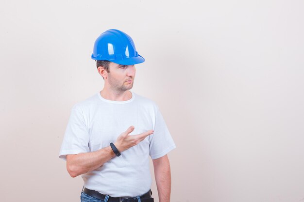 Trabajador de la construcción manteniendo la mano en gesto de desconcierto en camiseta, jeans, casco y mirando serio