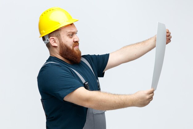 Trabajador de la construcción masculino joven concentrado con casco de seguridad y uniforme de pie en la vista de perfil estirando papel mirándolo aislado sobre fondo blanco