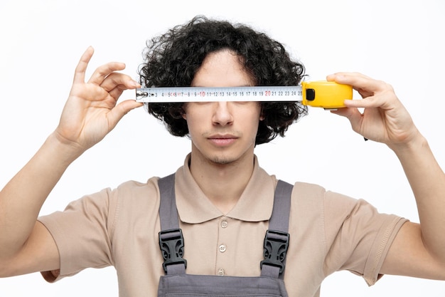 Foto gratuita trabajador de la construcción masculino joven serio vistiendo uniforme manteniendo la cinta métrica delante de los ojos aislado sobre fondo blanco.