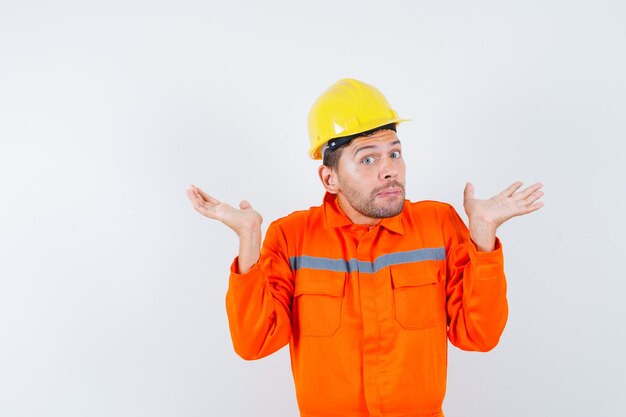 Trabajador de la construcción mostrando gesto de impotencia en uniforme, casco y mirando confundido, vista frontal.