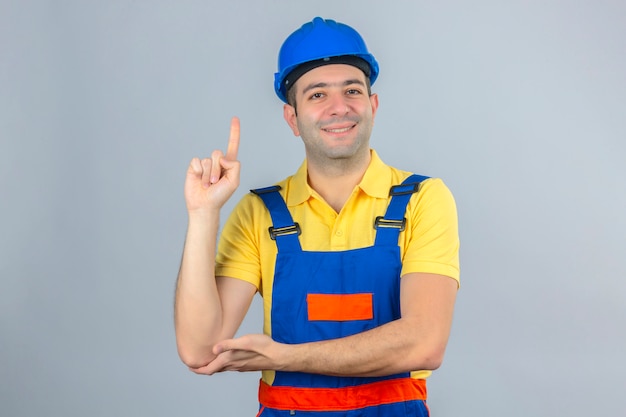 Trabajador de la construcción en uniforme y azul casco de seguridad apuntando con el dedo hacia arriba sonriendo positivo aislado en blanco