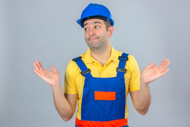 Trabajador de la construcción en uniforme y azul casco de seguridad despistado y expresión confusa con brazos y manos levantadas aislado en blanco