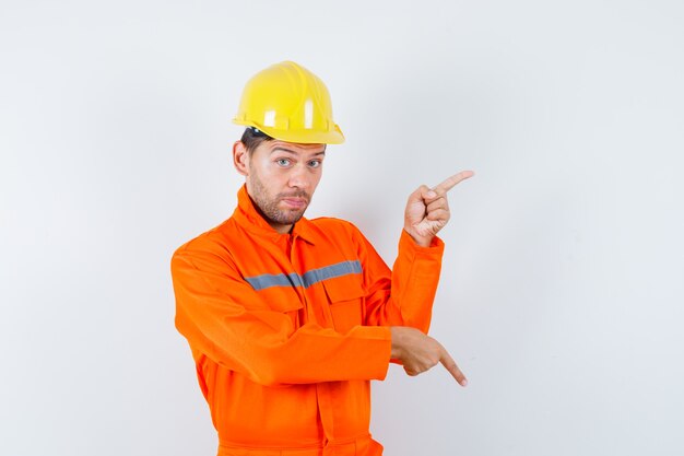 Trabajador de la construcción en uniforme, casco apuntando con el dedo hacia arriba y hacia abajo y mirando indeciso, vista frontal.