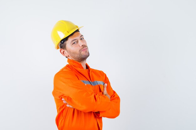 Trabajador de la construcción en uniforme, casco de pie con los brazos cruzados y mirando confiado, vista frontal.