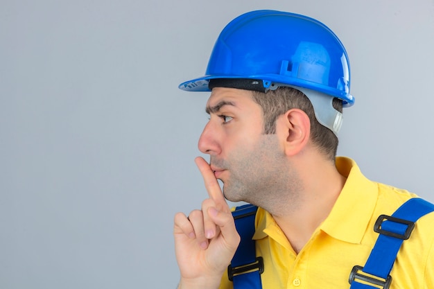 Trabajador de la construcción en uniforme y casco de seguridad azul haciendo gesto de silencio en blanco aislado