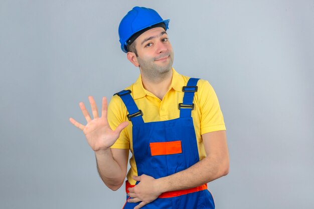 Trabajador de la construcción en uniforme y casco de seguridad azul mostrando y apuntando hacia arriba con los dedos número cinco con una sonrisa en la cara aislado en blanco