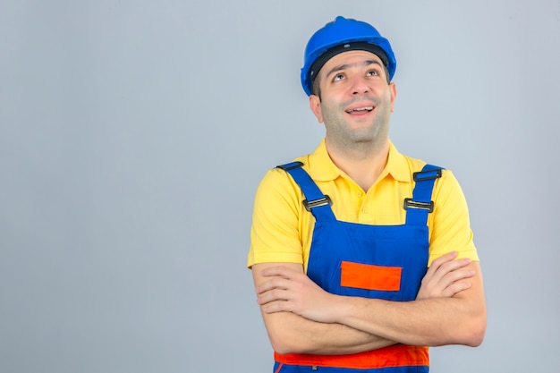 Trabajador de la construcción en uniforme y casco de seguridad azul pensando con una sonrisa en la cara y las manos cruzadas en blanco aislado