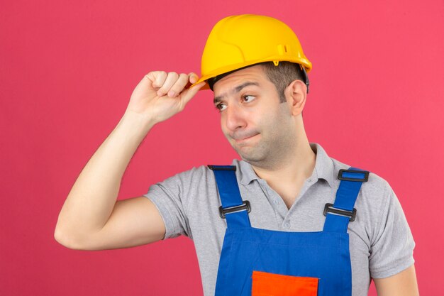 Trabajador de la construcción en uniforme y casco de seguridad con cara infeliz tocando su casco de seguridad amarillo aislado en rosa