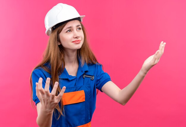 Trabajador constructor joven en uniforme de construcción y casco de seguridad mirando decepcionado levantando las manos sobre la pared rosa